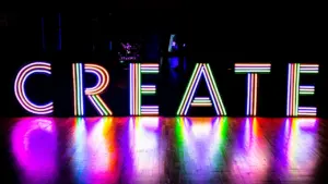Neon gaismas burti, kas veido vārdu Create, atrodas uz tumšas skatuves grīdas.