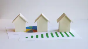Trīs koka mājiņas uz finanšu grafika, simbolizējot mājokļu kredītu un procentu likmju izmaiņas.