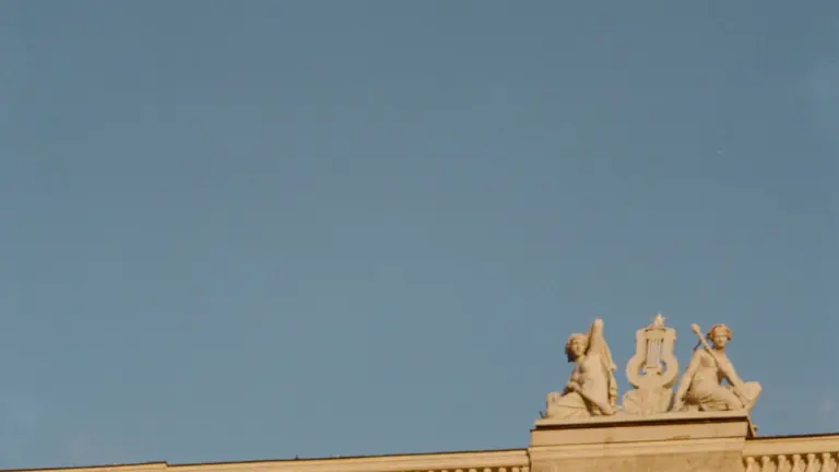 Latvijas Nacionālās operas ēkas fasādes fragments ar skulptūrām uz jumta gaišās debesīs.