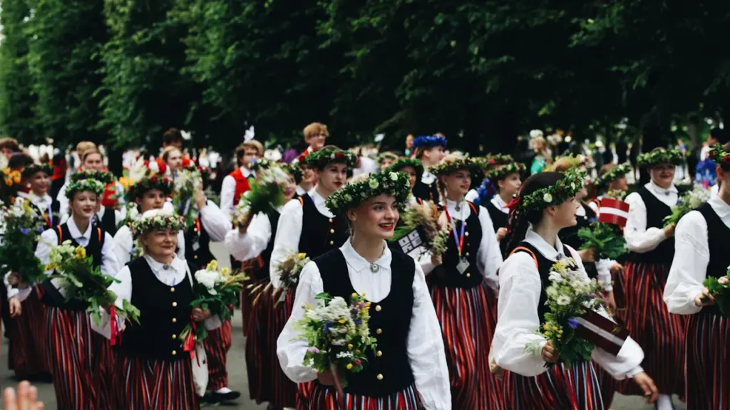 Grupa cilvēku latviešu tradicionālajos tautastērpos piedalās svinīgā gājienā ar ziediem rokās.