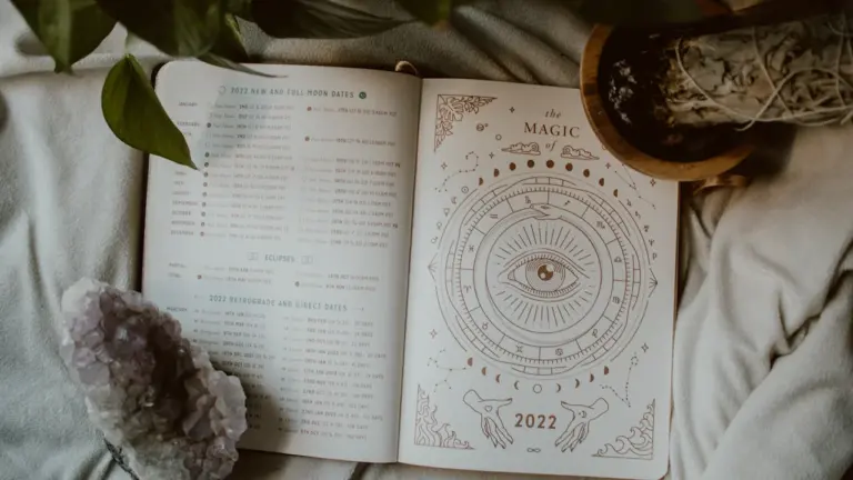 Atvērta grāmata ar astroloģijas simboliem uz gultas blakus zaļam telpaugam.