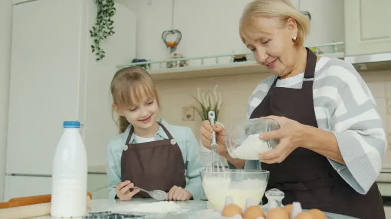 Vecmāmiņa māca mazmeitai gatavot mīklu mājīgā virtuvē, daloties ar ģimenes tradīcijām.