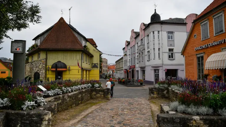 Vēsturiskā Cēsu pilsētas iela ar bruģakmeni, ziedu dobēm un tradicionālām Latvijas ēkām.