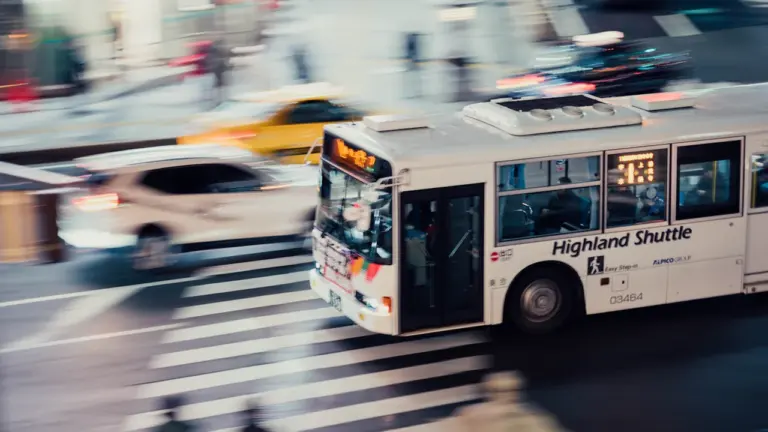 Autobuss brauc pa pilsētas ielu, simbolizējot steigu un nokavētu mirkli pilsētā.